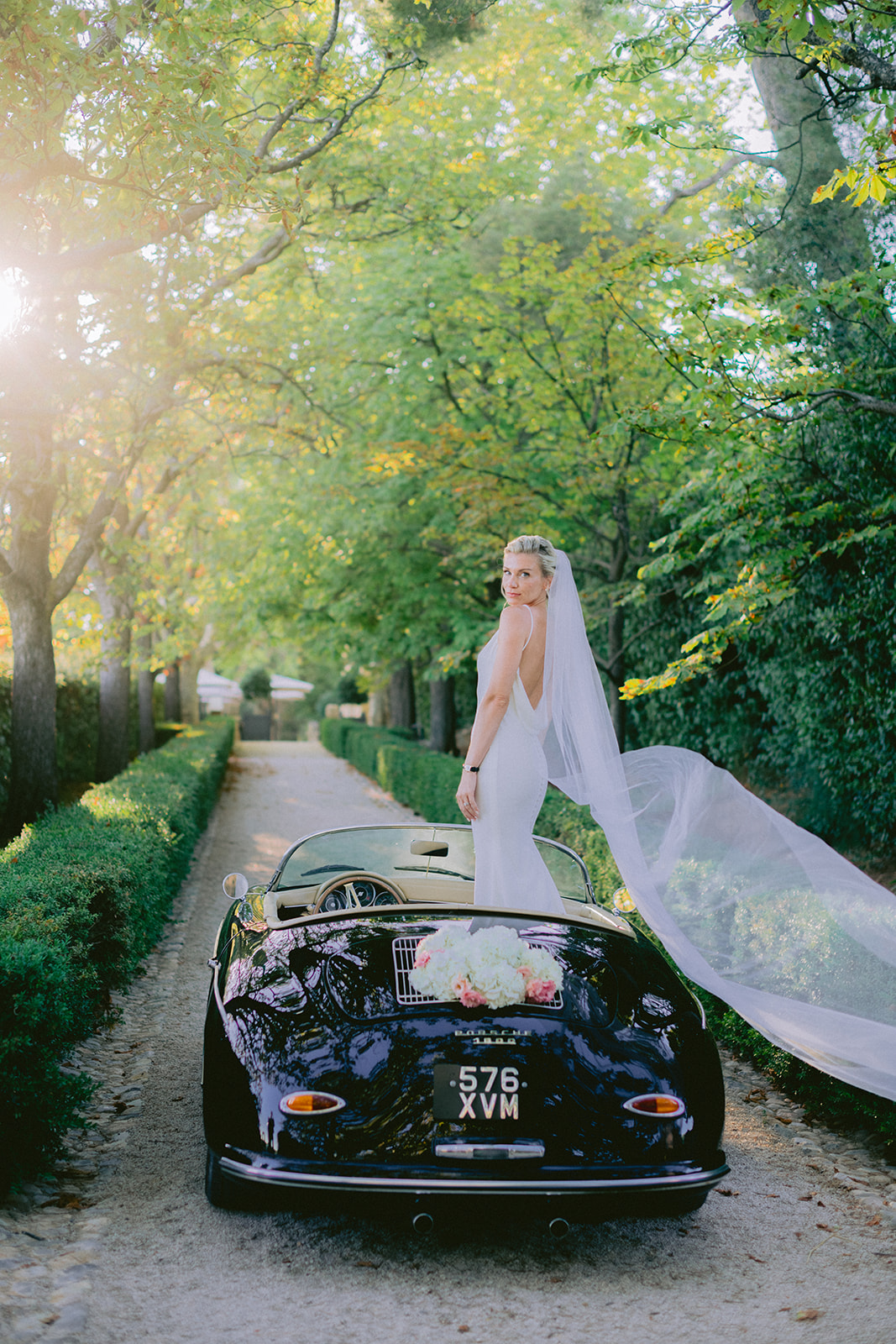 the bride is standing on the car and posing with her veil